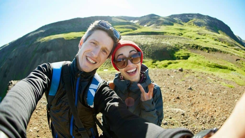 A couple smiles for a selfie on a sunny, mountainous landscape.