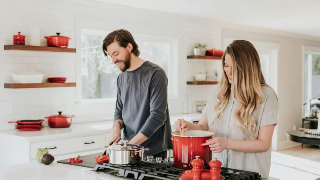 A smiling couple cooks together in a bright kitchen with red cookware.