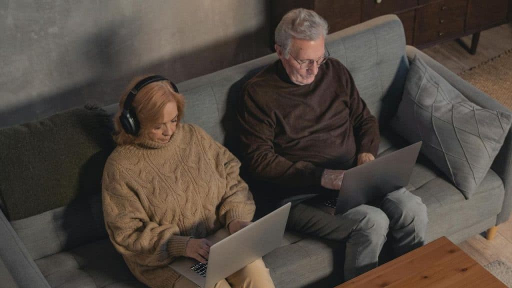 A man and a woman use laptops while sitting on a couch; the woman wears headphones.
