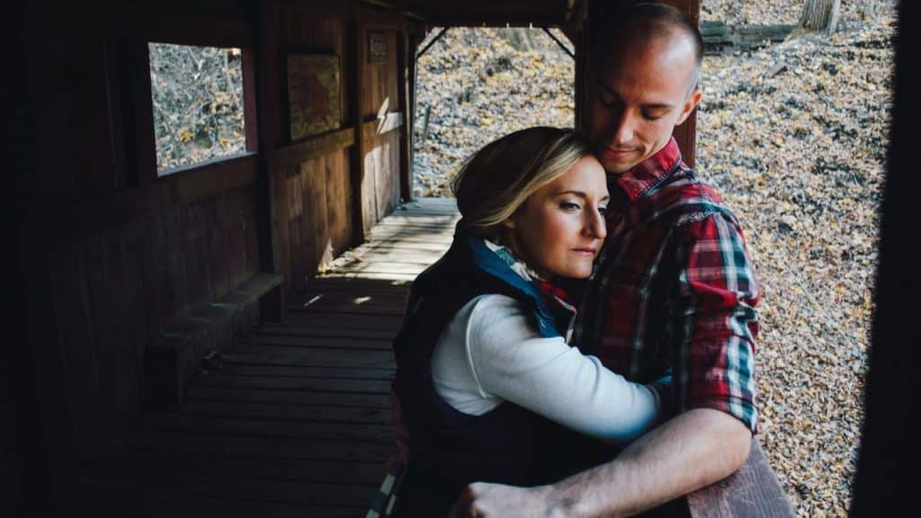 A man in a plaid shirt embraces a woman in a white top and navy vest on a wooden porch.