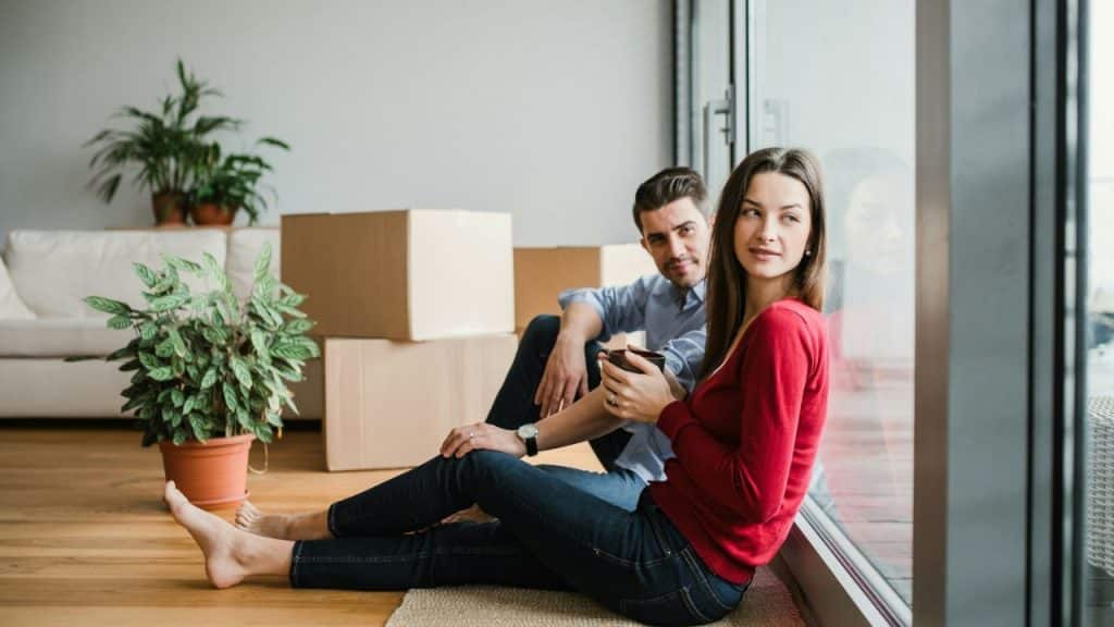 A woman holding a mug looks out a window while a man sits beside her, looking at the camera. Boxes are in the background.