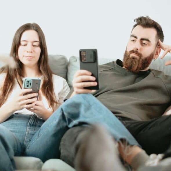 A man and a woman sit on a couch, both looking at their smartphones.