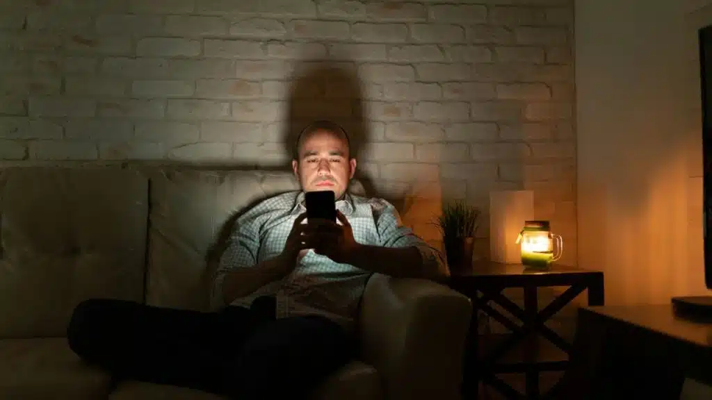 A man sits on a couch in a dimly lit room, illuminated by his phone.
