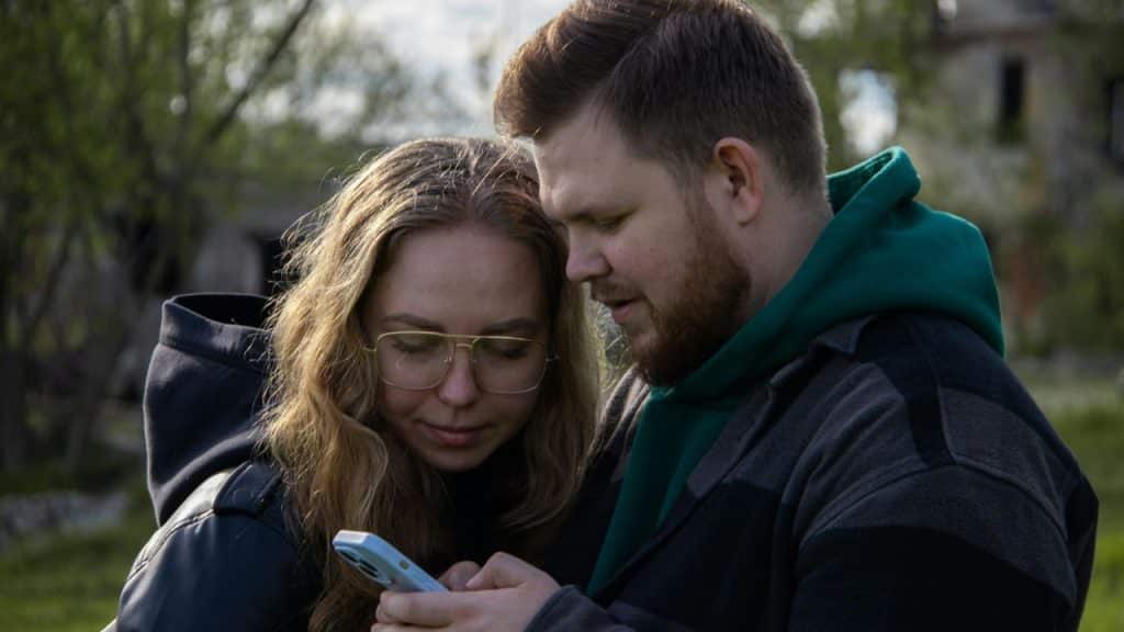 A man and a woman in glasses look closely at a smartphone outdoors.