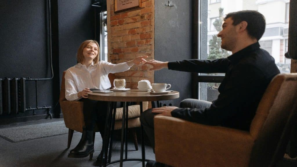 A smiling couple sits reaching out to touch hands at a cozy café table.