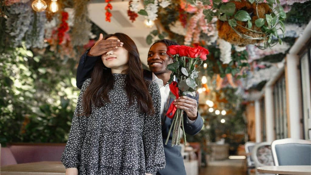 A man surprises a woman by covering her eyes while holding a bouquet of red roses