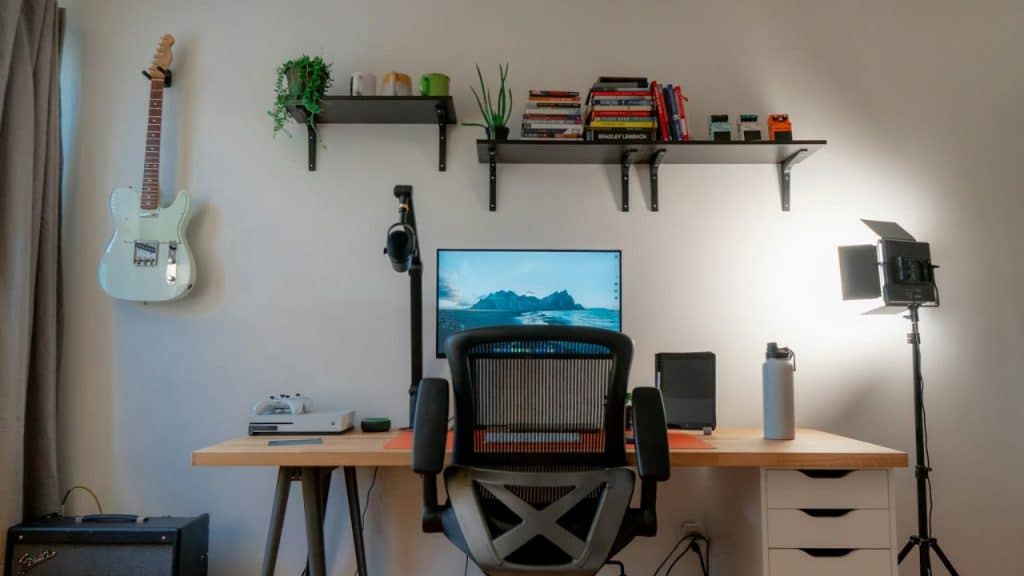 An ergonomic office setup features a desk with a monitor, chair, shelves, and a guitar on the wall.