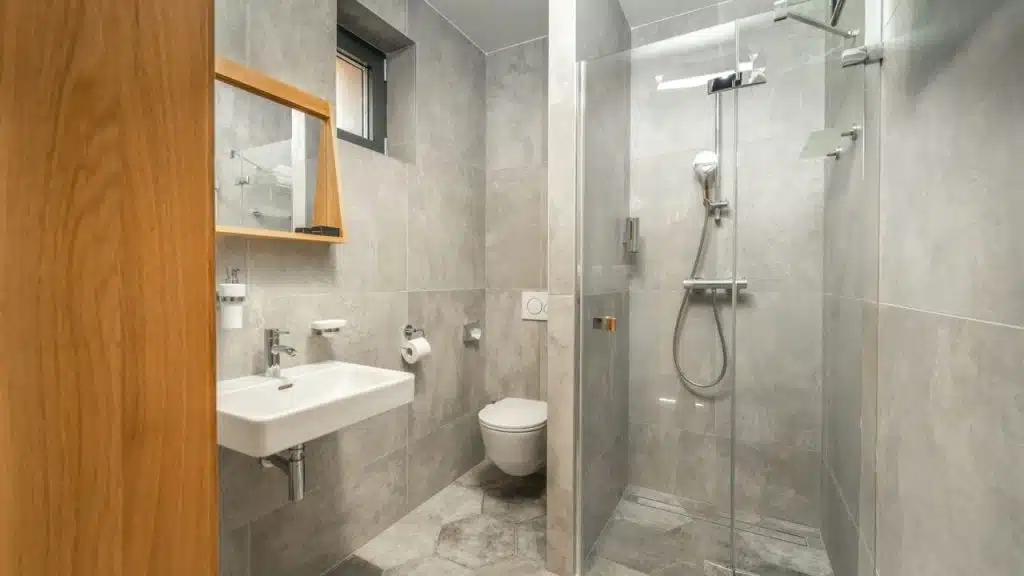 A modern bathroom with grey tiles features a shower, toilet, and sink with a wooden framed mirror.