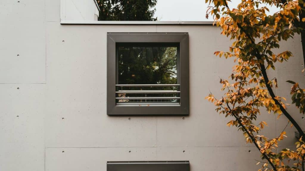 A modern building's concrete facade features a square window with horizontal bars and a tree with autumn leaves.