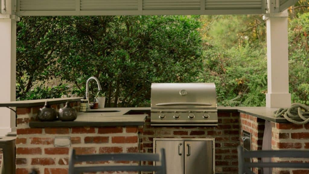 An outdoor kitchen features a built-in grill and sink on a brick base, surrounded by lush greenery.