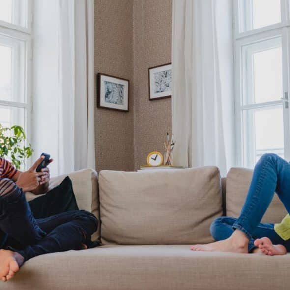 A man and a woman sit apart on a sofa, both looking at their phones, with windows behind them.