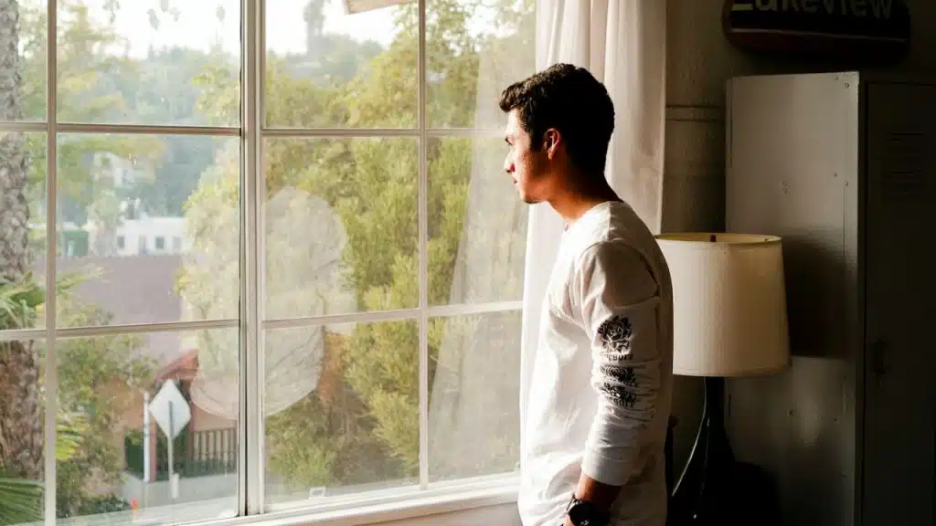 A man in a white long-sleeved shirt gazes out a large window, looking contemplative.