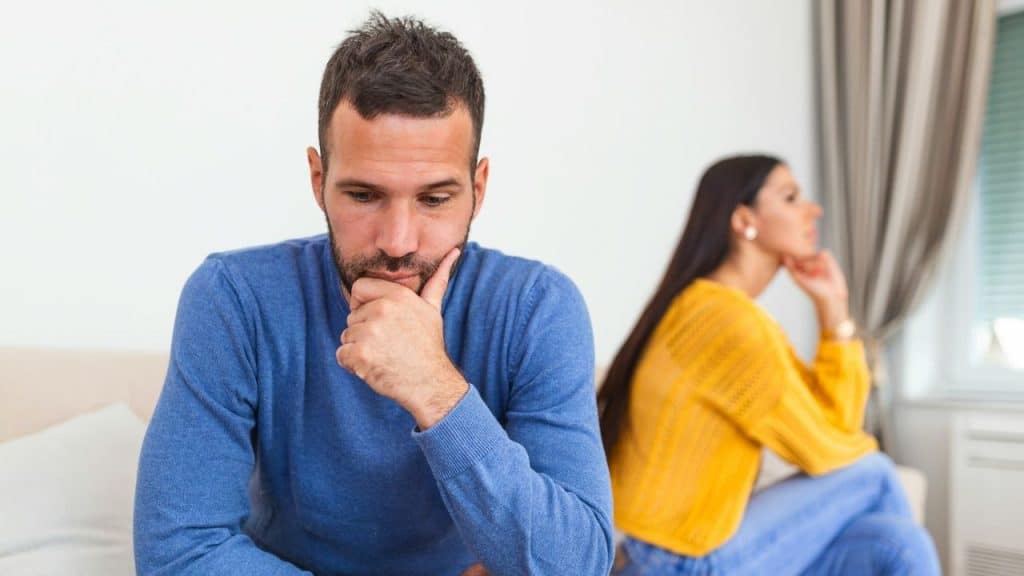 A thoughtful man in blue sits in the foreground, while a woman in yellow sits behind him, looking away.