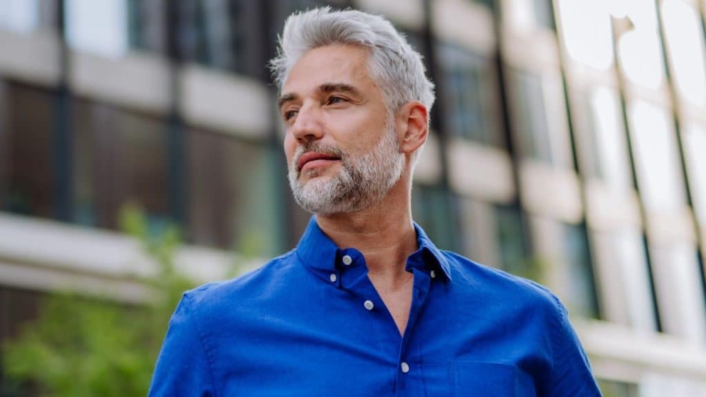 A handsome, gray-haired man in a blue shirt looks to his left, with a modern building in the background.