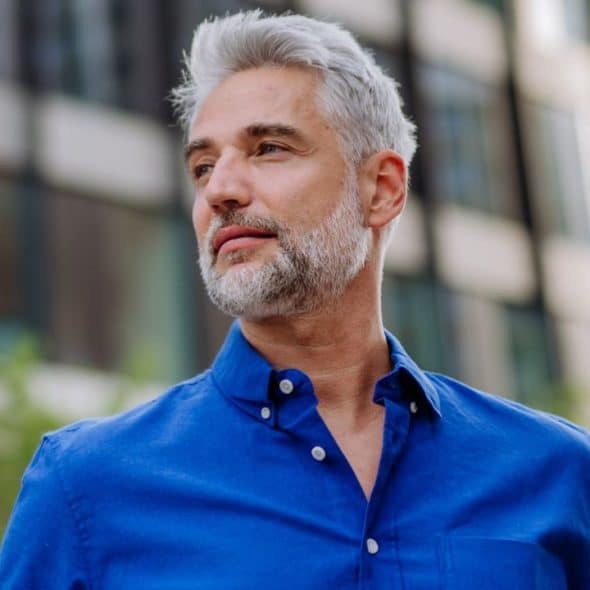 A handsome, gray-haired man in a blue shirt looks to his left, with a modern building in the background.