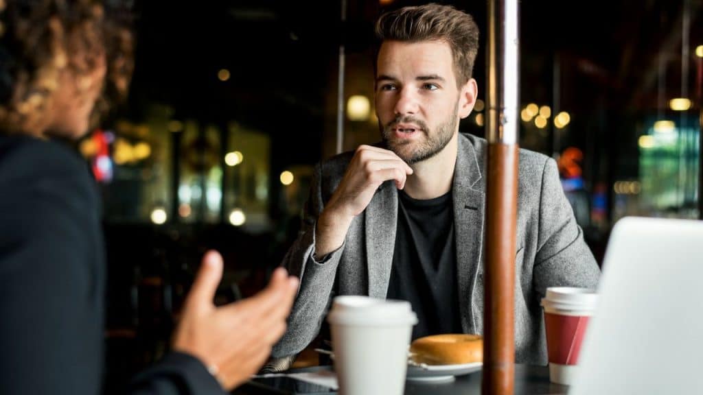 A man in a grey jacket listens intently to a person across the table.