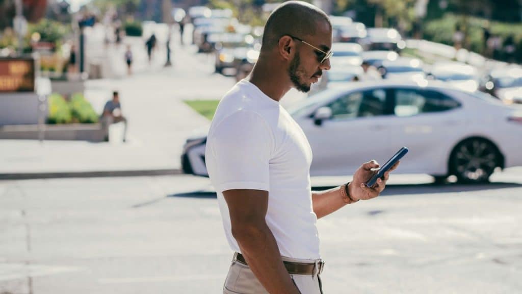 A man in a crisp white t-shirt and sunglasses looks at his phone on a sunny city street.