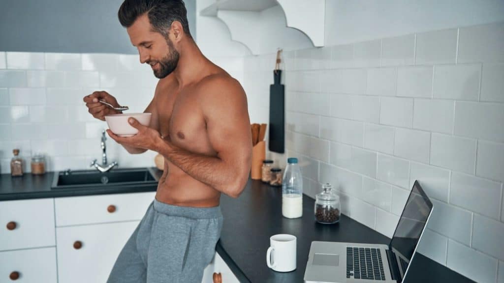 A shirtless man in gray sweatpants eats cereal in a modern kitchen with a laptop.