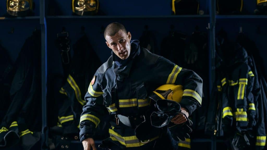 A male firefighter in uniform holds a helmet in a locker room.