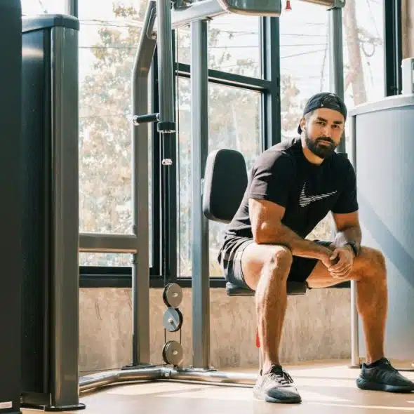A bearded man wearing a black shirt and cap sits on a gym machine, looking at the camera.