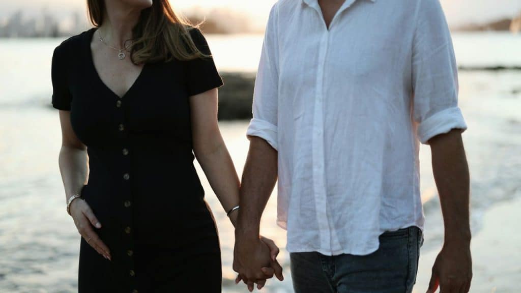 A middle-aged couple, a woman in a black dress and a man in a white shirt, hold hands outdoors.
