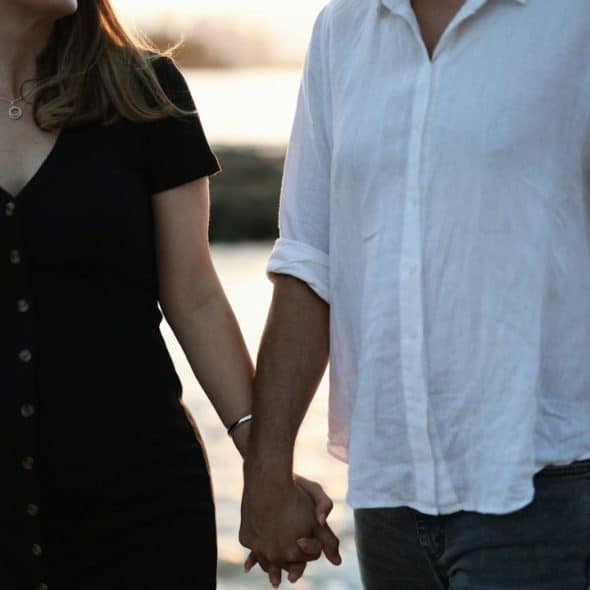 A middle-aged couple, a woman in a black dress and a man in a white shirt, hold hands outdoors.