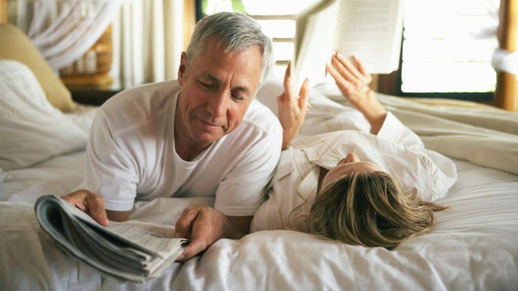 A mature couple lies in bed, a man reading a newspaper and a woman reading a book.