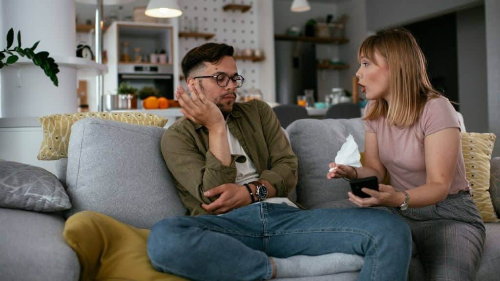 A man on a couch looks away as a woman, holding a phone and tissue, speaks to him.