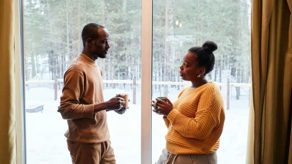 A man and woman, both holding mugs, stand by a window with a snowy outdoor view.