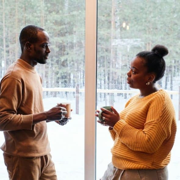 A man and woman, both holding mugs, stand by a window with a snowy outdoor view.
