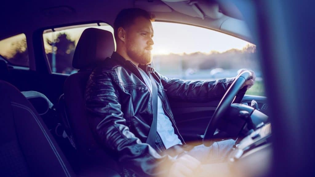 A man with a beard and a leather jacket holds a steering wheel with one hand.