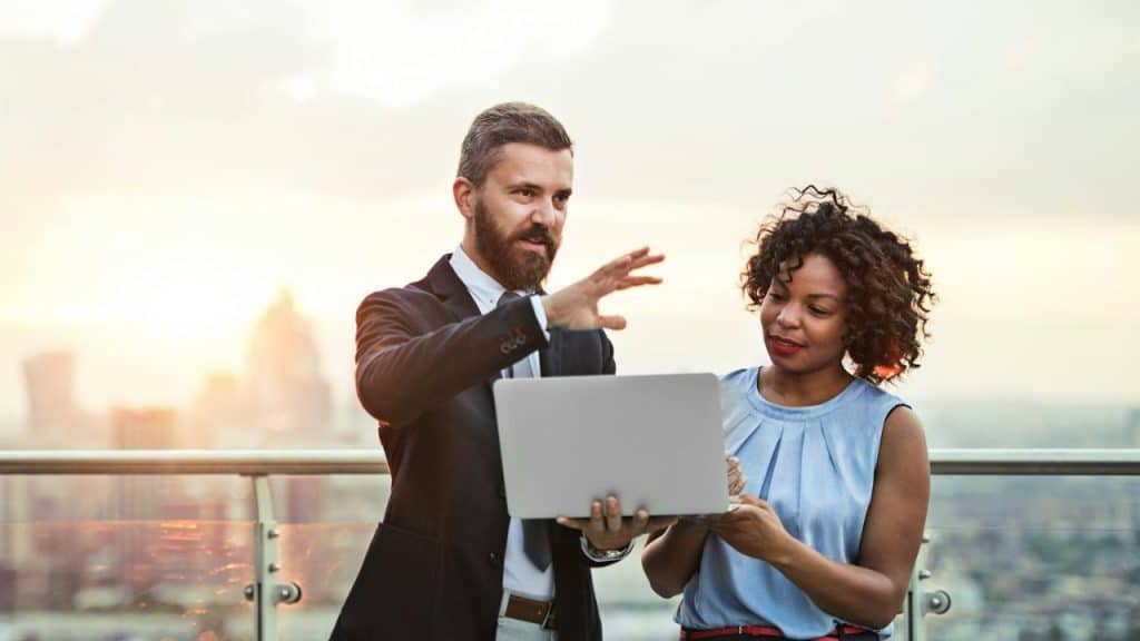 A man and woman discuss something on a laptop on a rooftop at sunset.