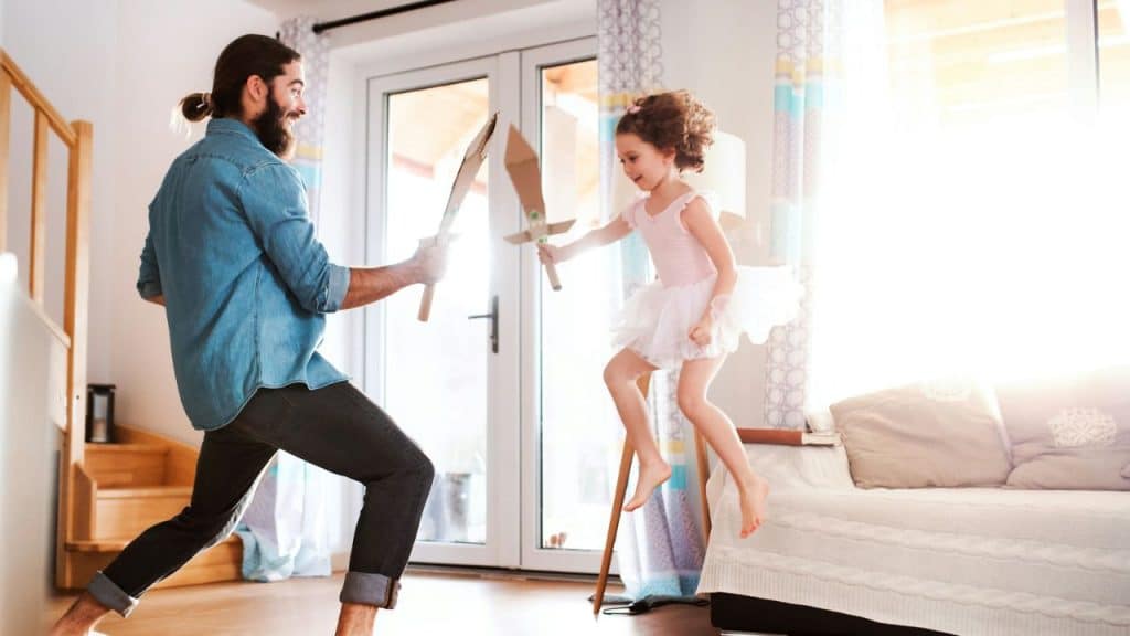 A man and a young girl play-fight with cardboard swords in a bright living room.