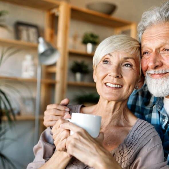 A happy elderly couple, with the man embracing the woman holding a mug, smiles in a cozy room.