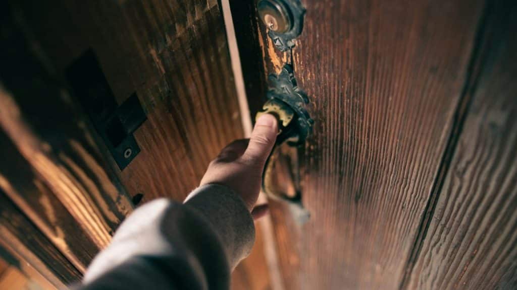 A hand is seen from a first-person perspective, knocking on an old, dark wooden door with a weathered doorknob and lock.