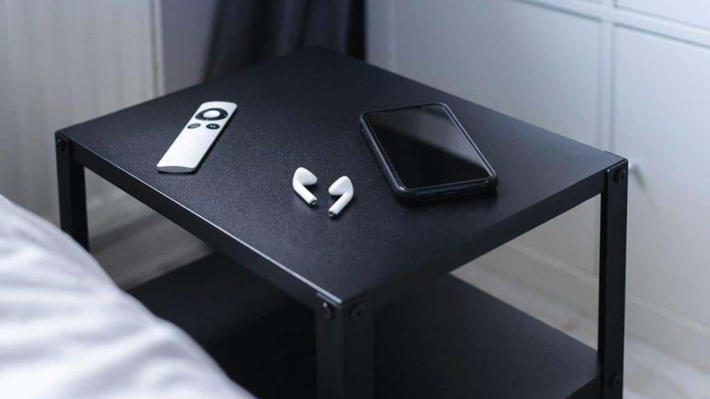 A white remote, wireless earbuds, and a black smartphone are on a black nightstand next to a bed.