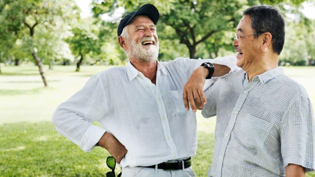 Two older men are outside, one with a beard and cap laughing, leaning on the other man.