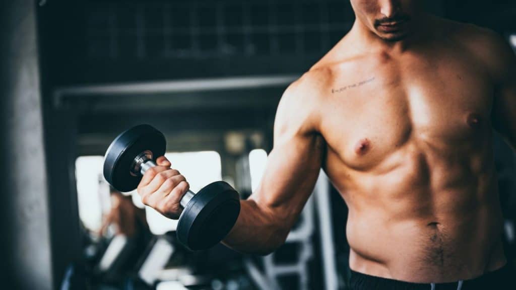 A shirtless, muscular man with a tattoo on his chest holds a dumbbell in a gym.