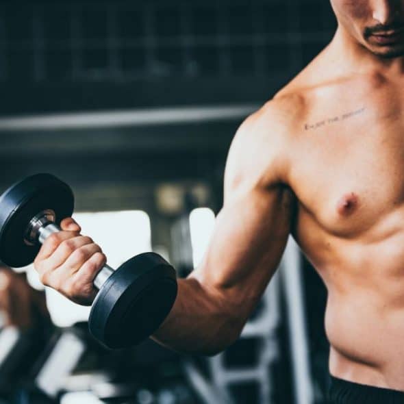 A shirtless, muscular man with a tattoo on his chest holds a dumbbell in a gym.