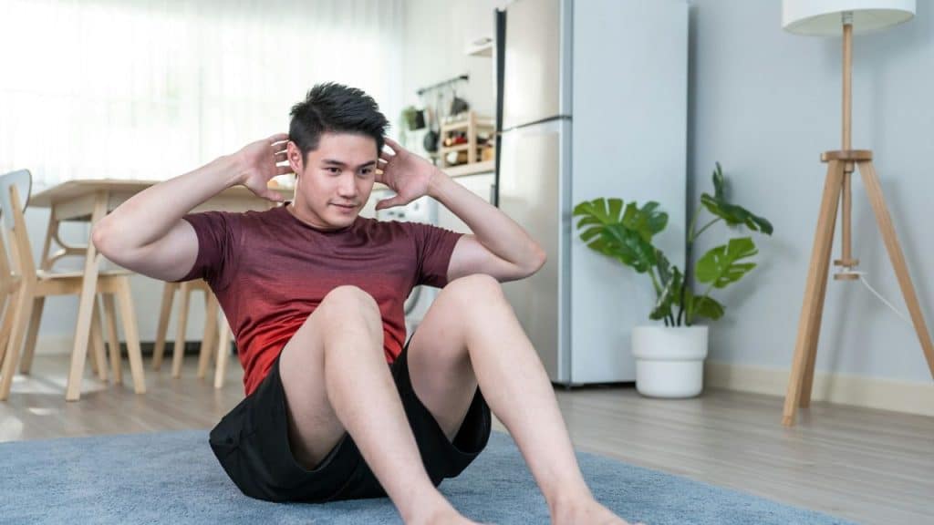 A man in a red shirt and black shorts does crunches on a mat in a living room.
