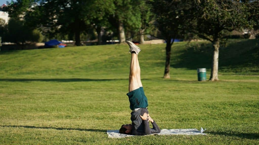 A person performs a shoulder stand yoga pose on a mat in a grassy park.