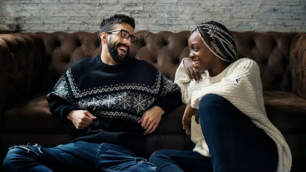 A smiling man and woman sit on the floor in front of a brown leather couch.