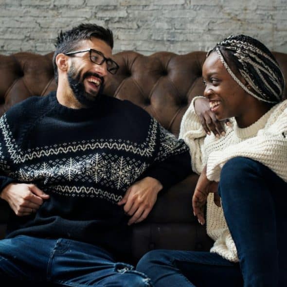 A smiling man and woman sit on the floor in front of a brown leather couch.