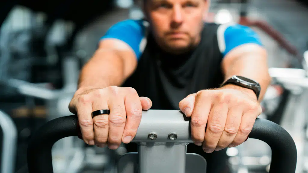 A man is gripping the handlebars of a fitness machine in a gym, wearing a smart ring on one hand and a smartwatch on the other.