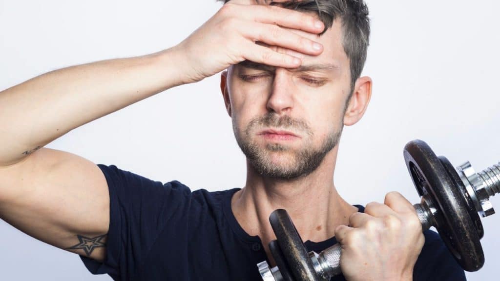 A man with messy hair holds a dumbbell and covers his forehead, looking exhausted.