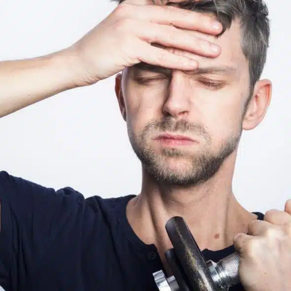 A man with messy hair holds a dumbbell and covers his forehead, looking exhausted.