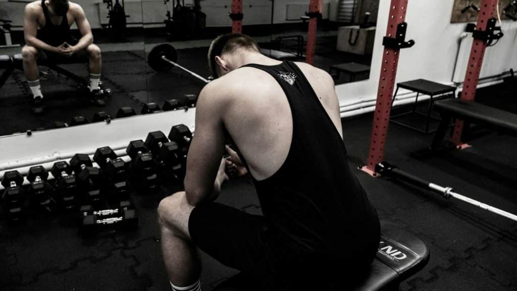 A person in a black tank top and shorts sits on a weight bench in a gym, facing away from the camera.