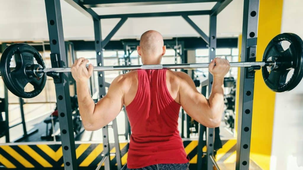 A bald man in a red tank top is seen from behind, holding a barbell on his shoulders under a squat rack.