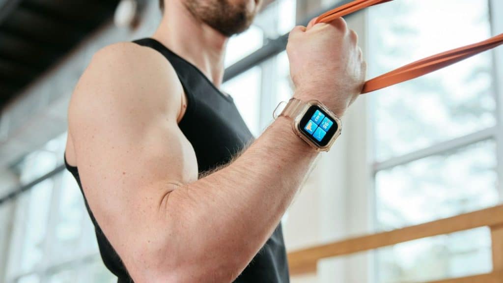 A man in a black tank top wears a smartwatch while holding an orange resistance band.