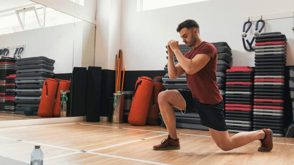 A man in a red shirt and black shorts performs a lunge in a gym with weights and mats.